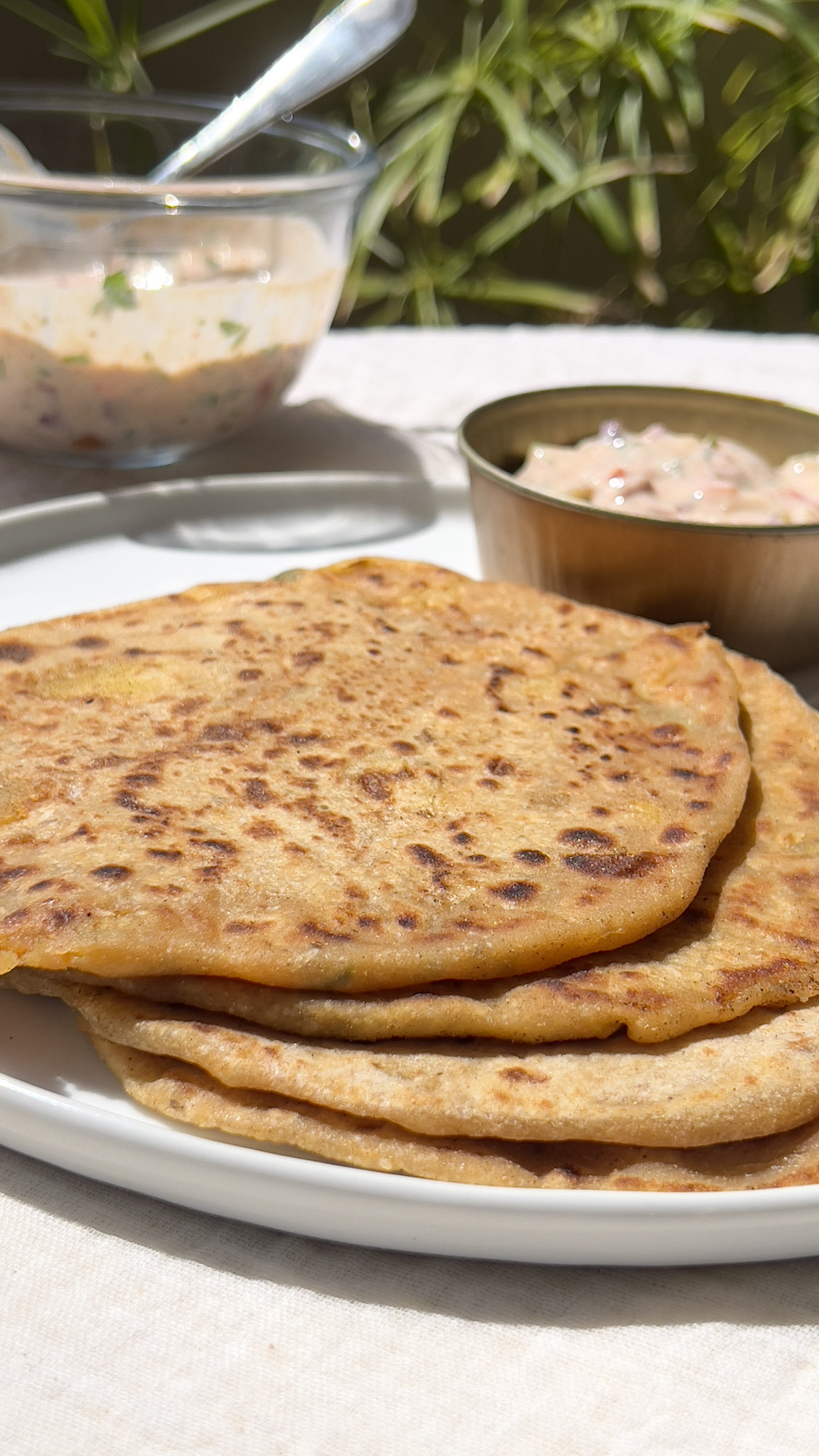 Aloo Parathas placed on a white plate with some raita in a bowl