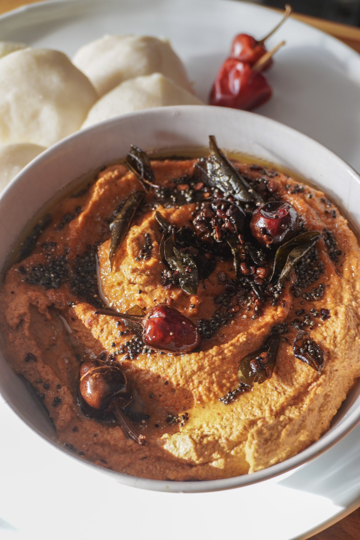 A close-up of a small bowl filled with vibrant red peanut chutney, topped with mustard seeds, curry leaves, and red chilies,