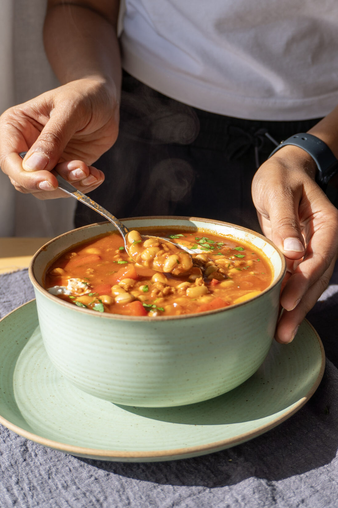 Front shot of chili garnished with cream and coriander, with me in the background holding a spoonful of the chili