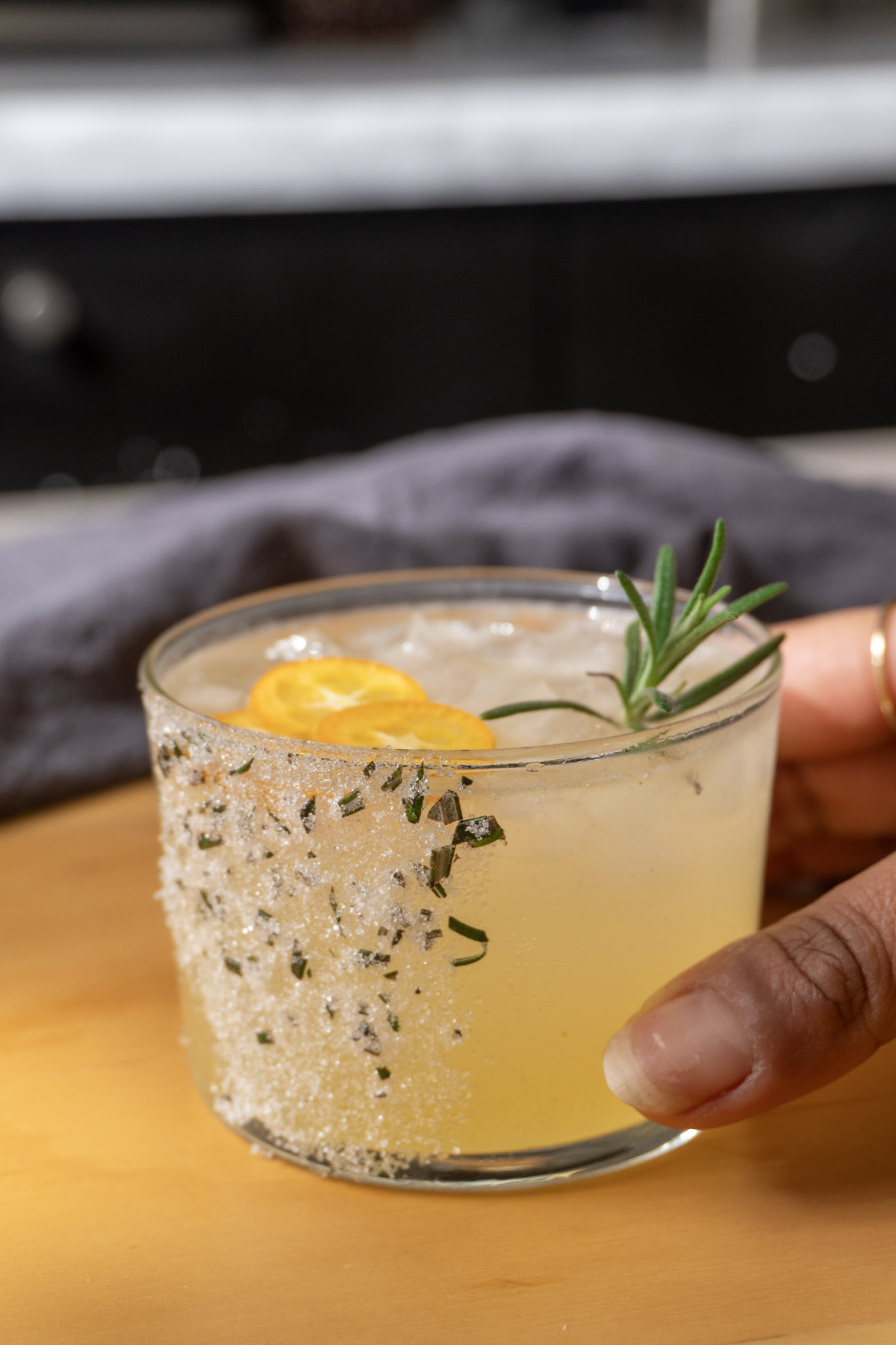 Holding up Kumquat Cardamom mocktail served in 1 rimmed glass with rosemary sprig as garnish.