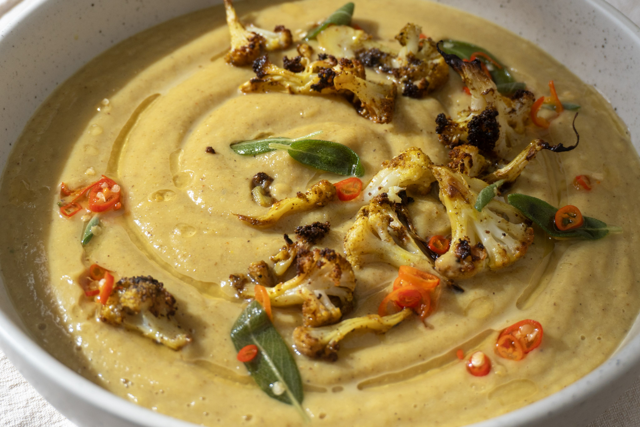Front shot of roasted cauliflower soup served in a large white bowl topped with toasted tofu and sage chili oil